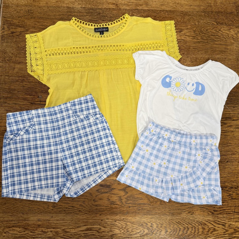 A yellow top with lace detailing, a white t-shirt with blue lettering that reads "GOOD things take time" and a flower graphic, paired with two sets of shorts: blue and white checked shorts and light blue shorts with daisy patterns, arranged on a wooden surface.