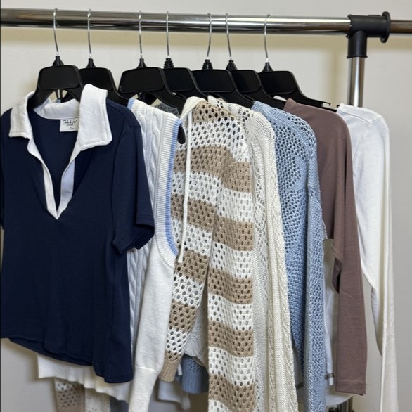 A collection of seven garments displayed on hangers, including a navy blue polo shirt with a white collar, a white sweater with a striped pattern, a light blue knit sweater, a beige cardigan, a white long-sleeve top, a light brown long-sleeve shirt, and a light gray long-sleeve top, arranged in a row against a neutral background.