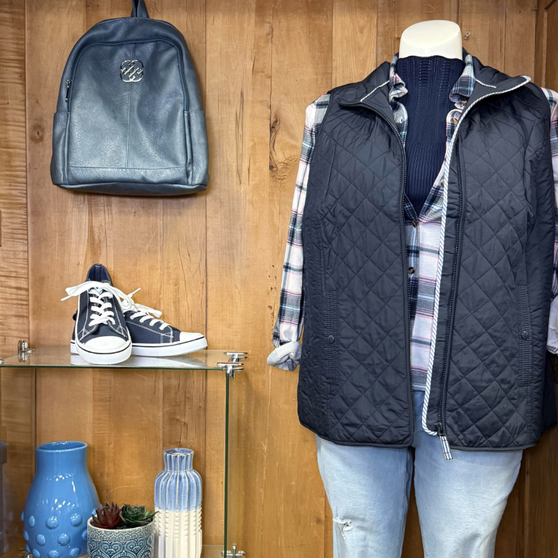 A display featuring a black quilted vest on a mannequin with a plaid shirt underneath, complemented by a navy blue knitted top. Below the mannequin, a pair of navy sneakers are placed on a glass shelf, alongside decorative items including a blue vase and a multi-striped container. A navy backpack hangs on the wooden wall in the background.