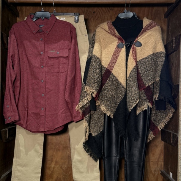 A red flannel shirt, a pair of beige pants, and a black poncho with checkered patterns hanging on hangers against a wooden background.